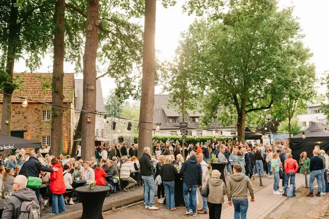Open-Air-Publikum an Stehtischen unter Bäumen im goldenen Abendlicht