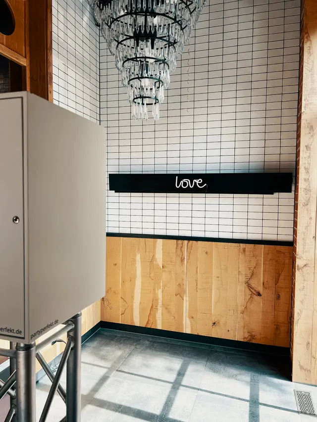 Fotobox vor einer weiß gefliesten Wand mit Love-Leuchtschrift und Kronleuchter